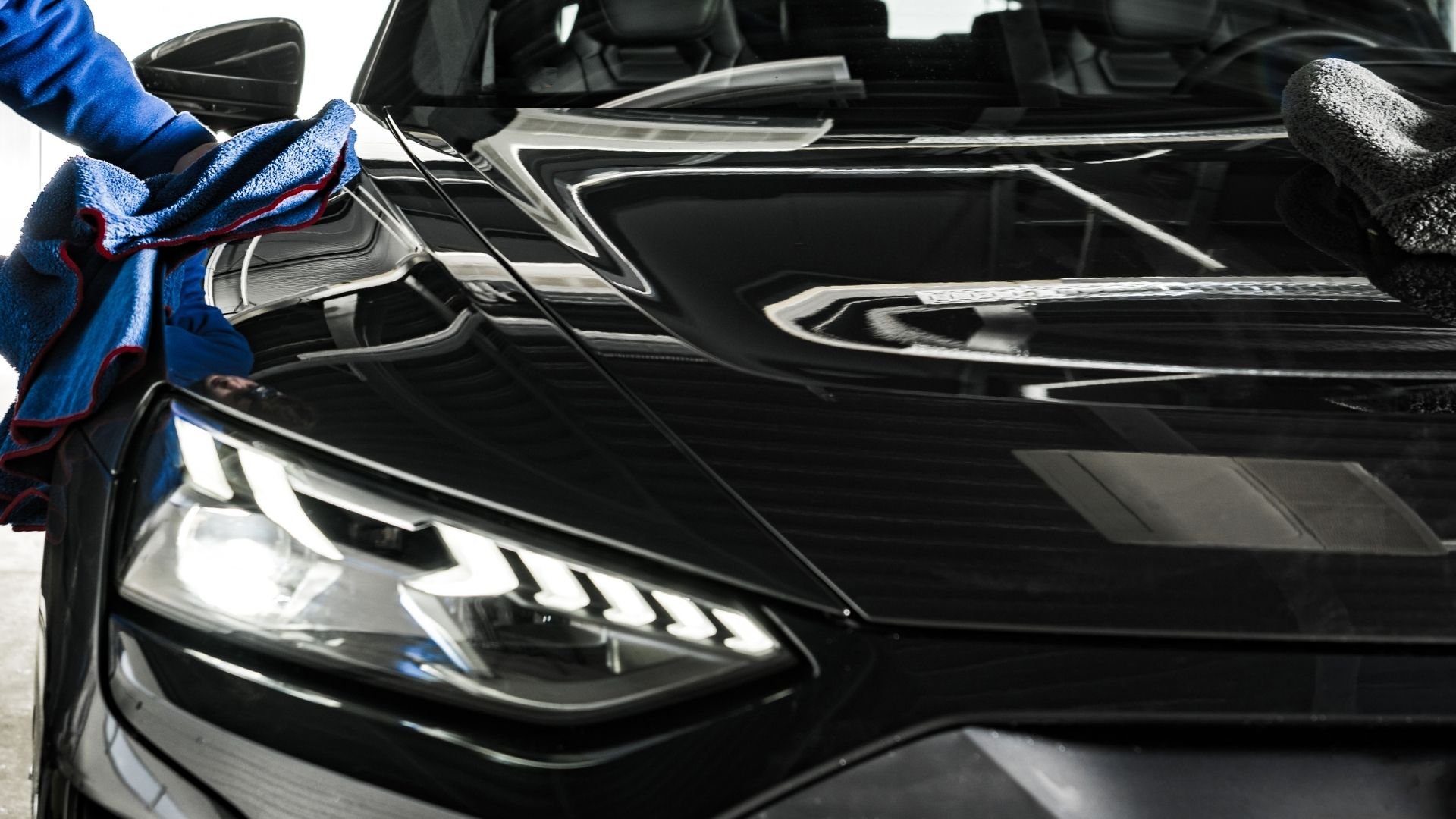 Person cleaning black car with blue microfiber cloth and squeegee tool