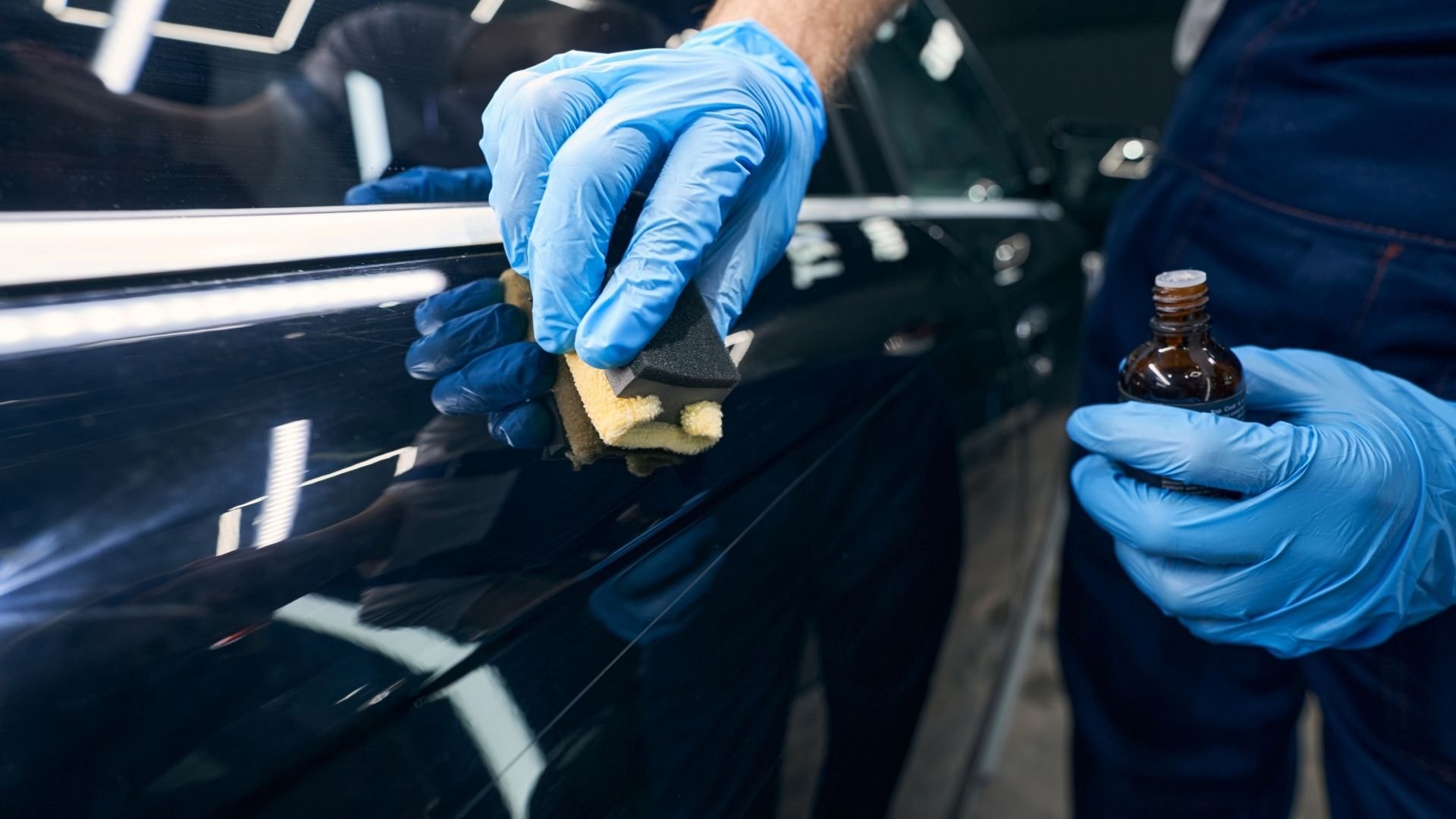 Technician in blue gloves applies protective coating to black car surface with applicator