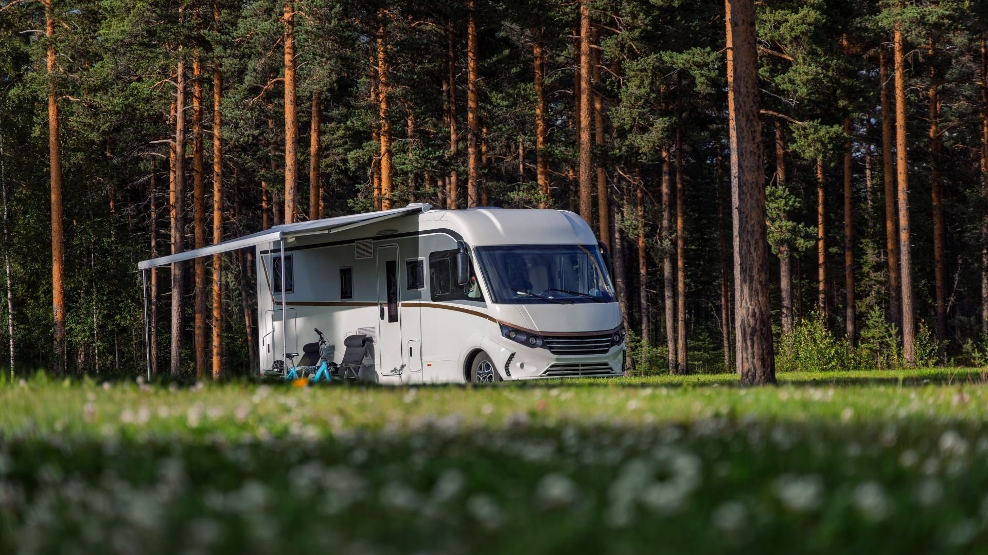 White modern motorhome with extended awning parked in forest clearing with tall pine trees.