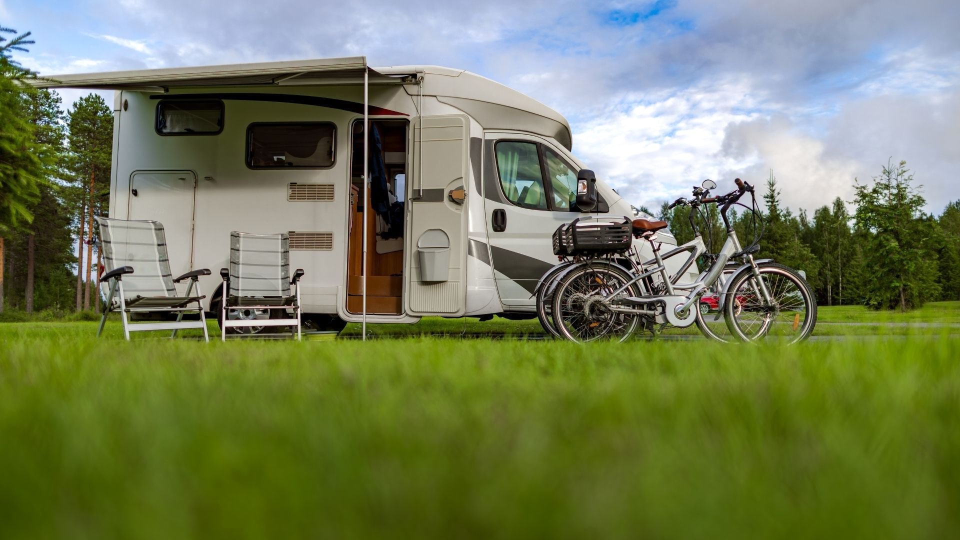 Motorhome with awning, camping chairs, and bicycles parked in grassy forest clearing