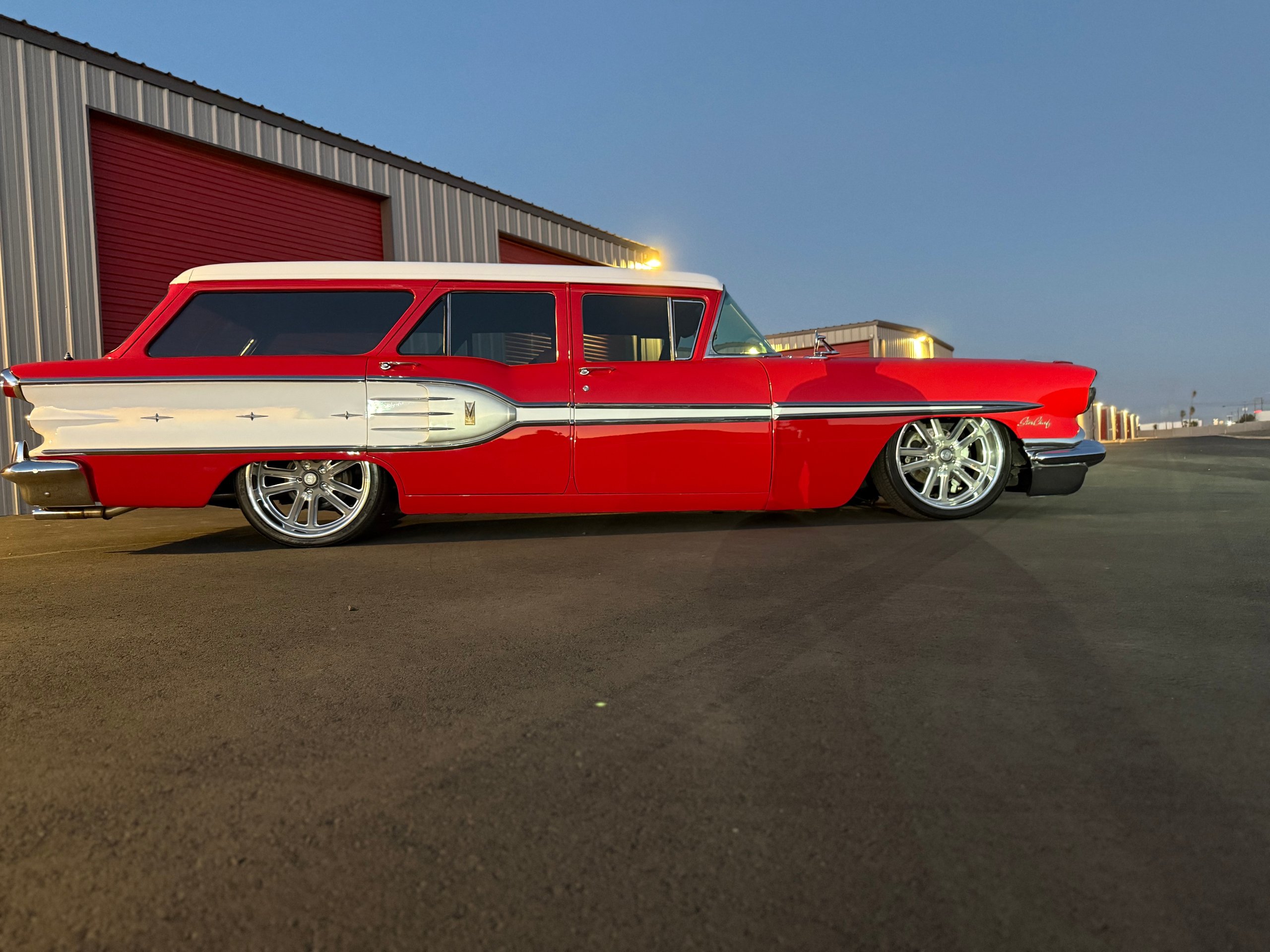 Red and white 1957 Chevrolet station wagon parked on asphalt outside industrial building